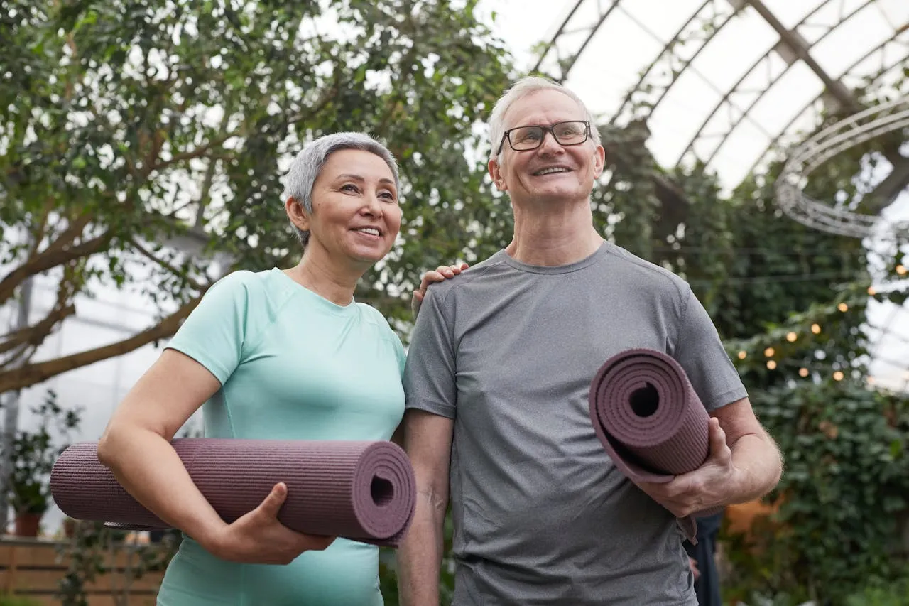 Smiling older couple standing in a garden, each holding a rolled-up exercise mat